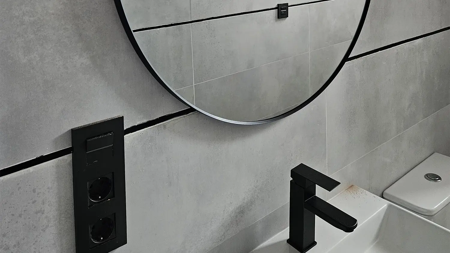 Baño moderno con espejo redondo, grifería negra y azulejos de gran formato en gris.