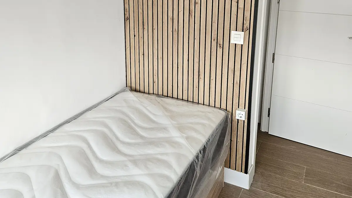 Cama individual con colchón nuevo junto a una pared decorada con listones de madera verticales.